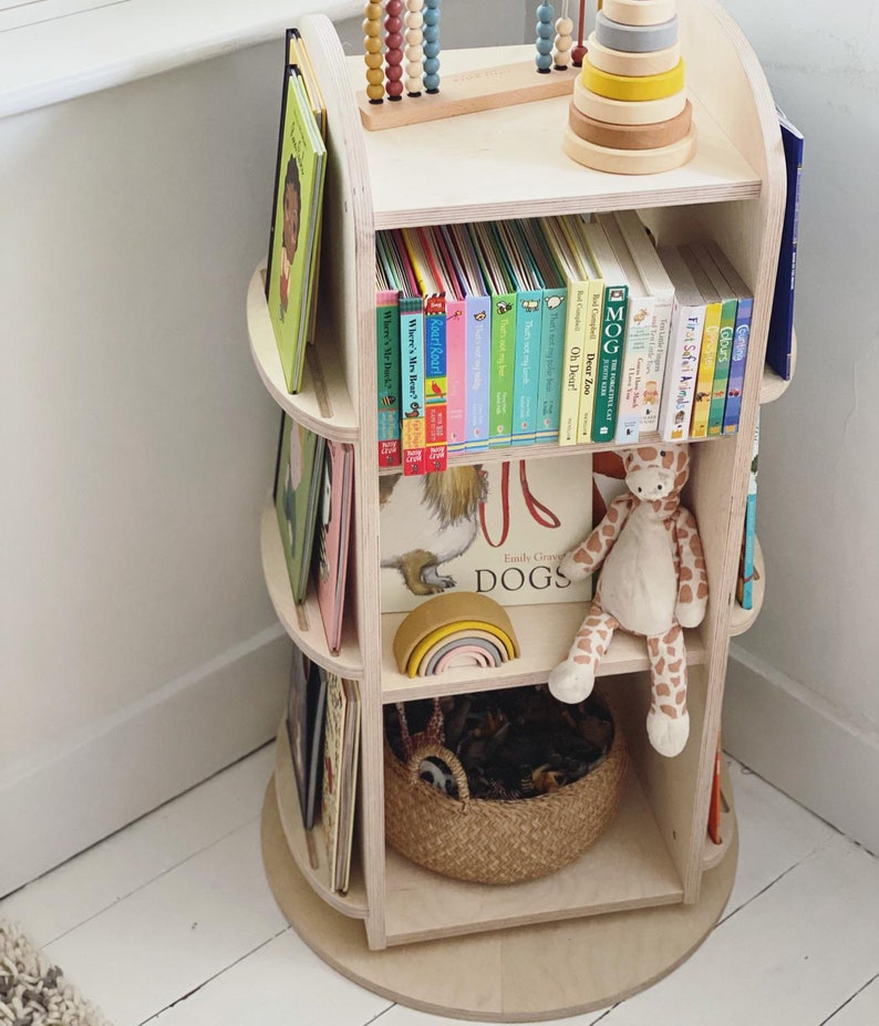 May include: A wooden rotating bookshelf with three shelves, filled with colorful children's books. The top shelf has a wooden abacus and a wooden stacking toy. The bottom shelf has a woven basket and a stuffed giraffe toy.