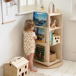 May include: A light wood children's bookshelf with rounded edges and a rotating base. The bookshelf is filled with colorful books and toys, including a stuffed giraffe, a wooden rainbow, and a wooden star. A young child is standing next to the bookshelf, reaching for a book.