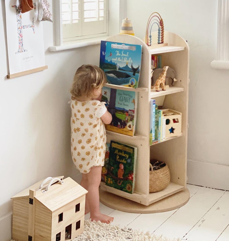 May include: A light wood children's bookshelf with rounded edges and a rotating base. The bookshelf is filled with colorful books and toys, including a stuffed giraffe, a wooden rainbow, and a wooden star. A young child is standing next to the bookshelf, reaching for a book.