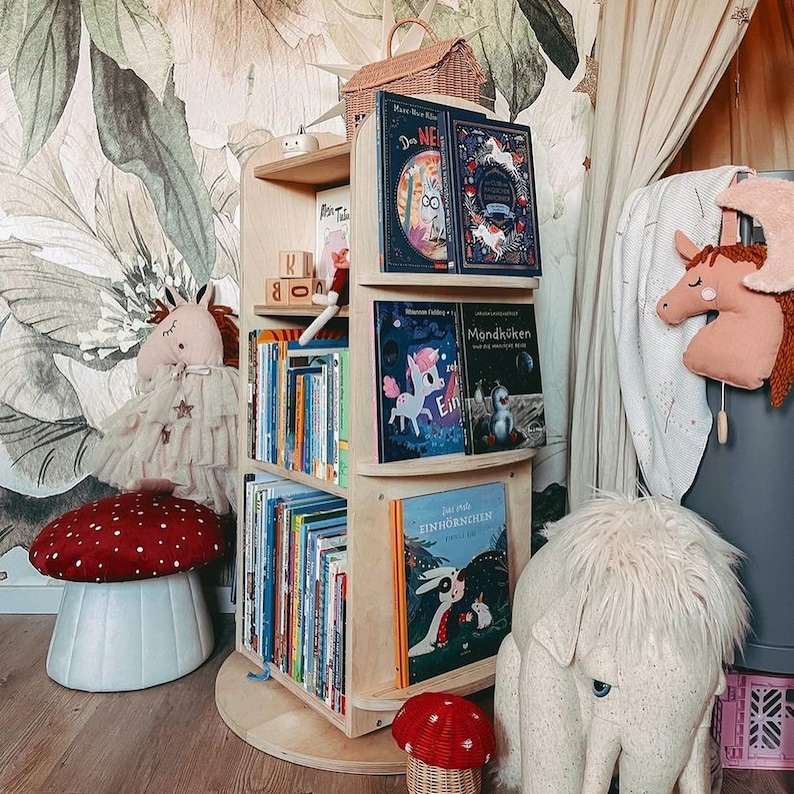 May include: A wooden bookshelf with five shelves, filled with children's books. The bookshelf is on a white base and has a white background. There are stuffed animals and a red mushroom stool in the foreground.