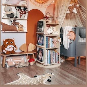May include: A nursery room with a white canopy bed, a wooden bookshelf filled with books, a wooden bench with a cat-shaped cushion, and a beige and brown animal-shaped rug. The walls are painted in shades of pink, orange, and white. The room is decorated with a star-shaped paper lantern and a mobile hanging above the bed.