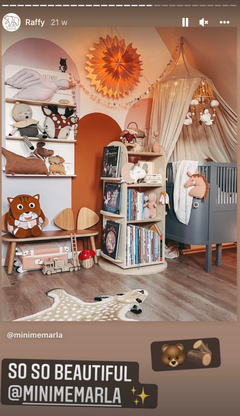 May include: A nursery room with a white canopy bed, a wooden bookshelf filled with books, a wooden bench with a cat-shaped cushion, and a beige and brown animal-shaped rug. The walls are painted in shades of pink, orange, and white. The room is decorated with a star-shaped paper lantern and a mobile hanging above the bed.