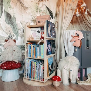 May include: A wooden rotating bookshelf with multiple shelves filled with colorful children's books. The bookshelf is in a nursery with a white and green floral wallpaper, a stuffed animal, and a crib.