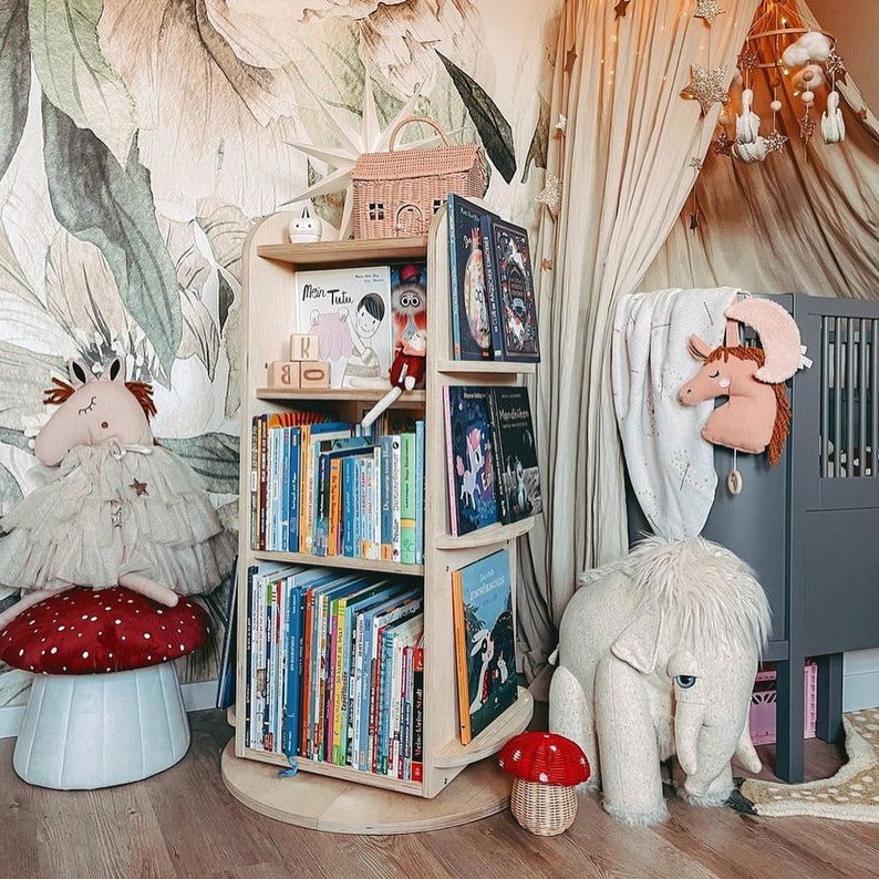 May include: A wooden rotating bookshelf with multiple shelves filled with colorful children's books. The bookshelf is in a nursery with a white and green floral wallpaper, a stuffed animal, and a crib.