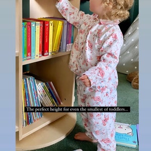 May include: A white wooden bookshelf with three shelves, filled with colorful children's books. A young child in a floral print pajamas reaches for a book on the top shelf. The text "The perfect height for even the smallest of toddlers..." is visible below the bookshelf. The text "Thanks for these lovely stories @ellescarboroughgooday" is visible at the bottom of the image.