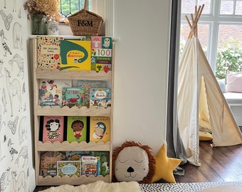 May include: A wooden bookshelf with a variety of children's books, including "I Had a Dinosaur", "100 First Words Nature", "Coco Chanel", "Seasid", and "Bizzy Bear". The bookshelf is in a playroom with a white teepee tent, a white fluffy rug, and a wooden balance board.
