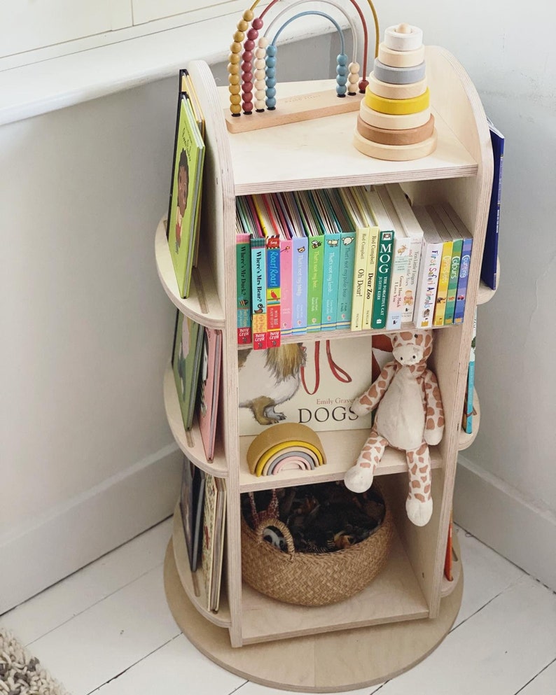 May include: A wooden rotating bookshelf with four shelves, filled with colorful children's books. The top shelf has a rainbow-colored wooden toy and a wooden stacking toy. A stuffed giraffe sits on the third shelf.