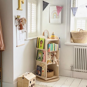 May include: A wooden rotating bookshelf with multiple shelves filled with children's books. The bookshelf is in a white room with a rug, a toy house, and a rainbow flag hanging on the wall.