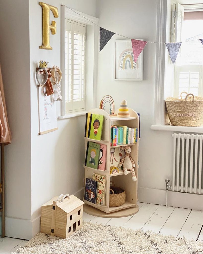 May include: A wooden rotating bookshelf with multiple shelves filled with children's books. The bookshelf is in a white room with a rug, a toy house, and a rainbow flag hanging on the wall.