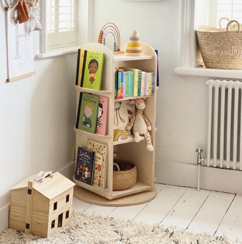 May include: A wooden rotating bookshelf with four shelves, filled with colorful children's books, a stuffed giraffe, and a rainbow wooden toy. The bookshelf is on a white rug in a room with white walls.