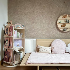 May include: A light pink bed with a wooden frame and storage compartments. The bed has a white and pink patterned pillow and a fluffy pink pillow. The wall behind the bed is covered in a brown wallpaper with a white floral pattern. A wooden bookshelf with books and toys is in the corner of the room.