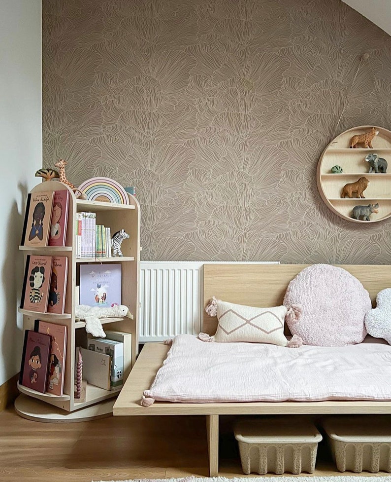 May include: A light pink bed with a wooden frame and storage compartments. The bed has a white and pink patterned pillow and a fluffy pink pillow. The wall behind the bed is covered in a brown wallpaper with a white floral pattern. A wooden bookshelf with books and toys is in the corner of the room.