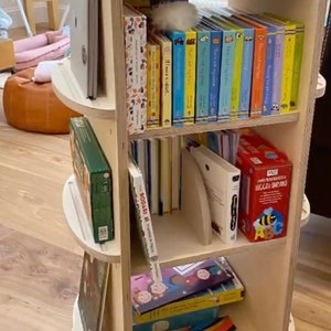 May include: A wooden rotating bookshelf with four shelves filled with colorful children's books. The top shelf has a wooden toy tree and a blue box with a white bird design. The bookshelf is on a wooden floor. The text "THANKS FOR SENDING THIS IN @THE_LITTLE_SUNBIRD" is at the bottom of the image.