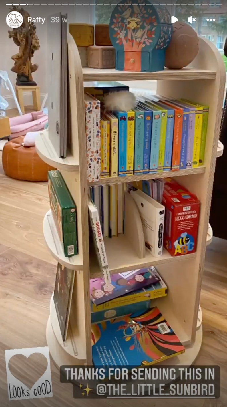 May include: A wooden rotating bookshelf with four shelves filled with colorful children's books. The top shelf has a wooden toy tree and a blue box with a white bird design. The bookshelf is on a wooden floor. The text "THANKS FOR SENDING THIS IN @THE_LITTLE_SUNBIRD" is at the bottom of the image.
