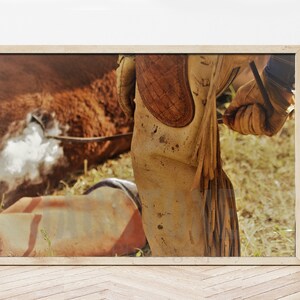 May include: Close-up of a cowboy boot with a brown leather shaft and a tan leather foot. The boot is worn and has dirt and mud on it. The boot is being worn by a cowboy who is standing in a field.
