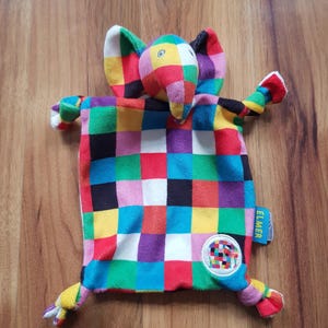 Może przedstawiać: Miękki pluszowy kocyk przytulanka z kolorową głową słonia w stylu patchwork, z dużymi uszami i trąbą. Kwadratowy korpus pokryty jest żywą kratką z czerwonych, niebieskich, żółtych, zielonych, fioletowych i białych kwadratów. Niebieska metka zawiera napis ELMER.