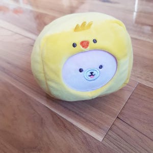Op de afbeelding: Een gele pluche speelgoed in de vorm van een kuiken, met een lichtroze gezicht en een lachend berengezicht. Het speelgoed heeft kleine zwarte ogen, een rode snavel en een kleine gele kam. Het ligt op een houten oppervlak.