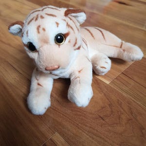 Pode incluir: Um brinquedo de pelúcia de filhote de tigre creme e castanho claro. O animal de pelúcia tem olhos grandes e expressivos, um nariz rosa e listras marrons. O brinquedo está deitado em uma superfície de madeira.