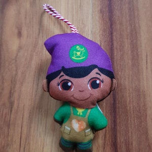 May include: Purple and green plush toy with a brown face, wearing a green shirt and brown pants. The toy has a green circle on its hat with a white reindeer inside.