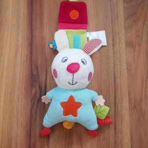 Puede incluir: Un juguete de peluche de conejo blanco con una estrella azul y naranja en el pecho. El conejo tiene una nariz roja y mejillas rosadas. El juguete está unido a un lazo colgante con una casa de tela multicolor.