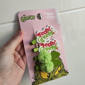 Könnte beinhalten: Ein Set Haarspangen im Grinch-Design auf einer rosa Karte. Die Karte zeigt das Gesicht des Grinch und die Worte "Feelin' Grinchy". Die Spangen umfassen grüne Pompons und grüne Spangen.