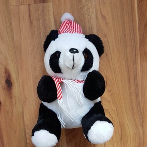 Puede incluir: Un oso panda de peluche con pelaje blanco y negro, que lleva un gorro y una bufanda a rayas rojas y blancas. El panda tiene orejas, ojos y patas negras. La bufanda es blanca con rayas rojas. El panda está sentado sobre una superficie de madera.