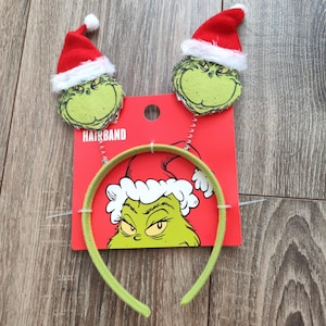 Könnte beinhalten: Ein grünes Stirnband mit zwei Grinch-Gesichtsdekorationen, jede mit einem roten Weihnachtsmannhut mit weißem Besatz. Das Stirnband befindet sich auf einer roten Karte mit dem Wort "HAIRBAND" und einer Grinch-Grafik.