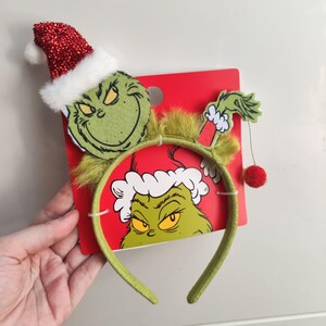 Könnte beinhalten: Ein festliches Stirnband mit einem grünen Grinch-Gesicht mit Weihnachtsmütze und rotem Bommel. Das Stirnband ist grün und hat ein rotes Etikett mit einem Grinch-Bild. Ideal für Weihnachten oder Feiertagsfeiern.
