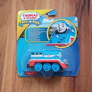 May include: A blue and red Thomas & Friends Take-n-Play toy train engine with a number 1 on the side. The train is on a blue and white cardboard package with the words "Thomas & Friends Take-n-Play" on the front.