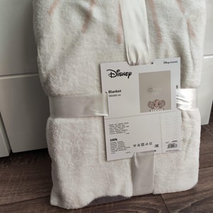 Puede incluir: Manta de forro polar blanca con un patrón rosa y blanco. La manta está doblada y tiene una etiqueta adjunta que dice "Disney" y "Blanket 150x160 cm". La etiqueta también tiene una imagen de un cachorro de león.