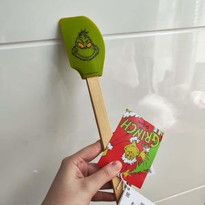 Könnte beinhalten: Ein grüner Silikon-Teigschaber mit dem Gesicht des Grinch auf dem Kopf und einem Holzgriff. Der Teigschaber wird von einem Etikett mit der Figur des Grinch und dem Text "Dr. Seuss" begleitet.