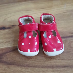Pu&ograve; includere: Un paio di scarpe rosse per bambini piccoli con teste di Topolino bianche stampate su di esse. Le scarpe hanno un cinturino bianco e una suola bianca.