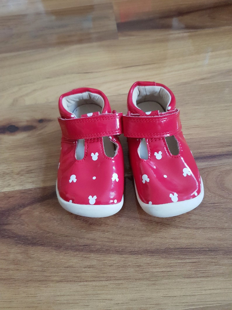 Pu&ograve; includere: Un paio di scarpe rosse per bambini piccoli con teste di Topolino bianche stampate su di esse. Le scarpe hanno un cinturino bianco e una suola bianca.