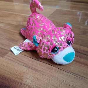 Peut inclure: Léopard en peluche rose et or avec des yeux bleus et un nez turquoise. Le léopard est couché sur le ventre, la queue enroulée.