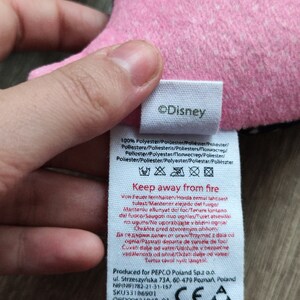 K&ouml;nnte beinhalten: Ein wei&szlig;es Etikett mit schwarzem Text, auf dem "&copy;Disney" und "Keep away from fire" steht. Das Etikett enth&auml;lt auch Pflegehinweise und Informationen &uuml;ber die Herkunft und den Hersteller des Produkts. Das Etikett ist an einem rosa und schwarzen Stoff befestigt.