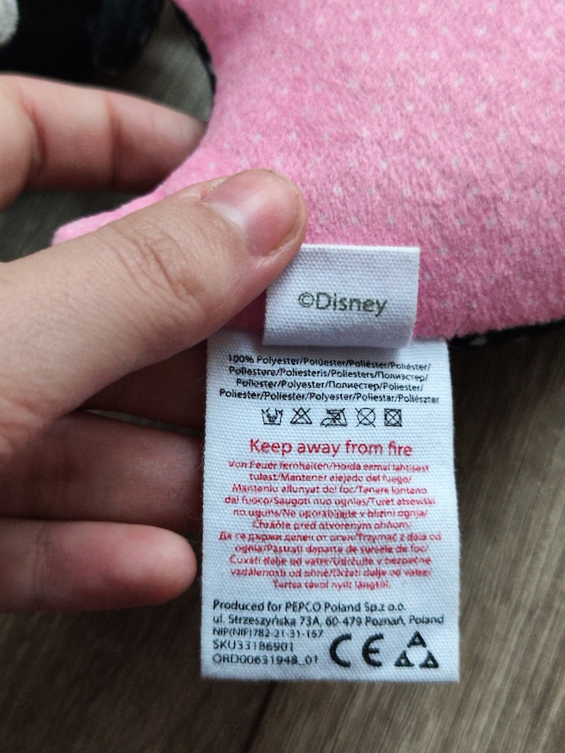 K&ouml;nnte beinhalten: Ein wei&szlig;es Etikett mit schwarzem Text, auf dem "&copy;Disney" und "Keep away from fire" steht. Das Etikett enth&auml;lt auch Pflegehinweise und Informationen &uuml;ber die Herkunft und den Hersteller des Produkts. Das Etikett ist an einem rosa und schwarzen Stoff befestigt.