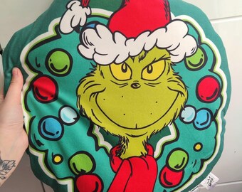 Peut inclure: Un coussin vert sur le thème du Grinch en forme de couronne. Le Grinch porte un bonnet et une écharpe rouges de Père Noël, avec une couronne d'ornements colorés autour de son visage. Une étiquette est attachée.