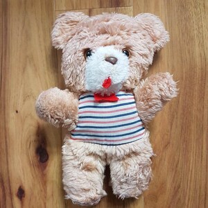 May include: A plush teddy bear with light brown fur, a white muzzle, and a red bow tie. The bear wears a striped shirt with red, white, and blue horizontal stripes. The bear's arms are outstretched.