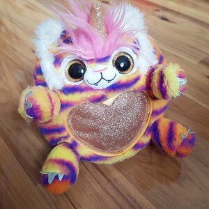 Puede incluir: Un peluche con forma de híbrido tigre-unicornio. El juguete presenta un cuerpo a rayas amarillas, naranjas y moradas, ojos negros grandes y una melena rosa. También tiene un cuerno dorado con purpurina y un corazón con purpurina.