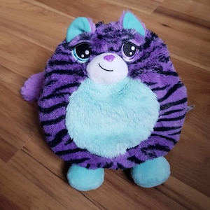 Pode incluir: Um brinquedo de pelúcia redondo em forma de gato com pelo roxo e listras pretas. O gato tem barriga, orelhas e pés azul claro. Possui olhos grandes e um pequeno nariz roxo.
