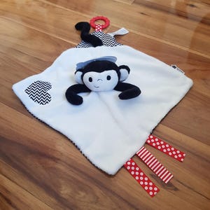 Op de afbeelding: Een wit en zwart chevron-patroon babydeken met een zwart en wit apen gezicht in het midden. De deken heeft een rood en wit stippenlint en een rood en wit gestreept lint bevestigd aan de onderste hoek. De aap heeft een zwarte staart met een rood en wit ringetje bevestigd aan het einde.