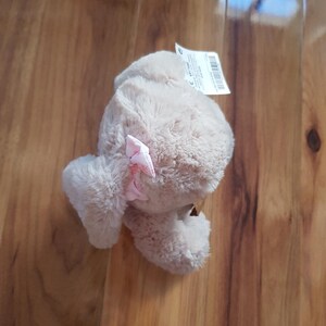 May include: A beige plush toy with a pink bow. The soft toy has a furry texture and is lying on a wooden surface. A white tag with text is attached to the toy.