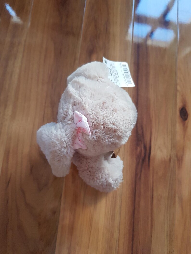 May include: A beige plush toy with a pink bow. The soft toy has a furry texture and is lying on a wooden surface. A white tag with text is attached to the toy.