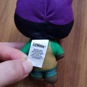 May include: A plush toy elf wearing a purple and black hat, green shirt, brown pants, and green and purple striped socks. The toy has a tag that reads "Elf Mates /TM/&copy; 2022 CCA and B, LLC dba The Lumistella Company. All Rights Reserved. Licensed by Rocket Licensing Ltd."