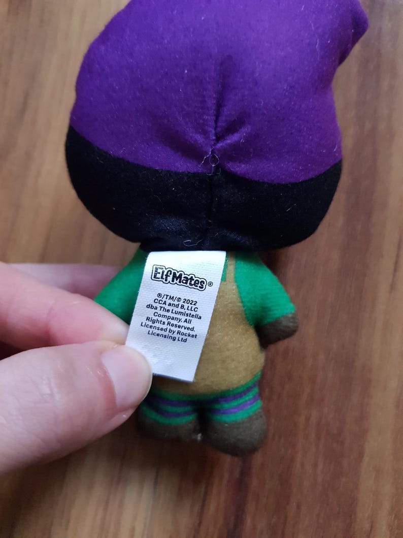May include: A plush toy elf wearing a purple and black hat, green shirt, brown pants, and green and purple striped socks. The toy has a tag that reads "Elf Mates /TM/&copy; 2022 CCA and B, LLC dba The Lumistella Company. All Rights Reserved. Licensed by Rocket Licensing Ltd."