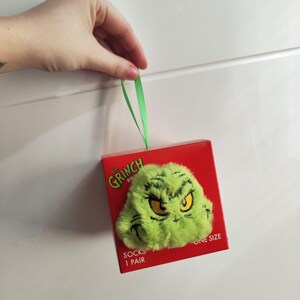 Peut inclure: Une boîte rouge avec un visage vert et poilu du Grinch aux yeux jaunes. La boîte porte l'inscription "The Grinch" et "Socks 1 Pair". Un ruban vert est attaché à la boîte.