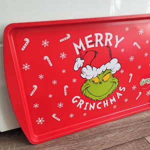 Op de afbeelding: Een rechthoekig rood dienblad met afgeronde hoeken. De dienblad toont de Grinch met een kerstmuts, met de woorden "MERRY GRINCHMAS" in het wit. Witte sneeuwvlokken en zuurstokken versieren de rode achtergrond.
