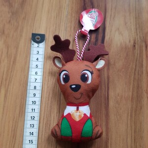May include: A plush reindeer ornament with a red and green outfit, a gold star on its chest, and a red tag that says "Reindeer".