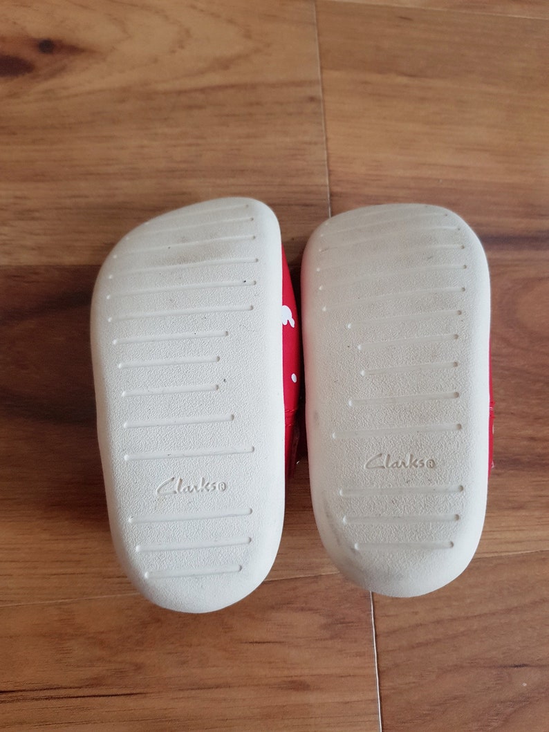 Pu&ograve; includere: Un paio di scarpe rosse per bambini piccoli con pois bianchi. Le scarpe sono della marca Clarks e hanno suole bianche.