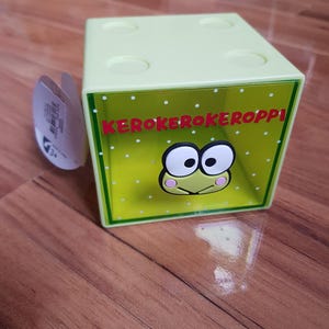 May include: A light green cube-shaped box with the words "KEROKEROKEROPPI" in red letters. The box features a cartoon frog face with large eyes and pink cheeks. The box has a clear plastic front and a light green top with four circular cutouts.